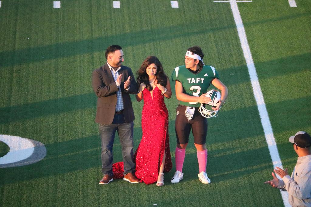 Two individuals and a woman in a red dress pose together on a football field, showcasing a vibrant outdoor setting.