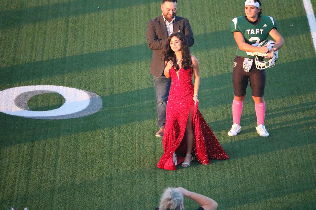 A woman in a red dress stands confidently on a football field, surrounded by green grass and goalposts in the background.