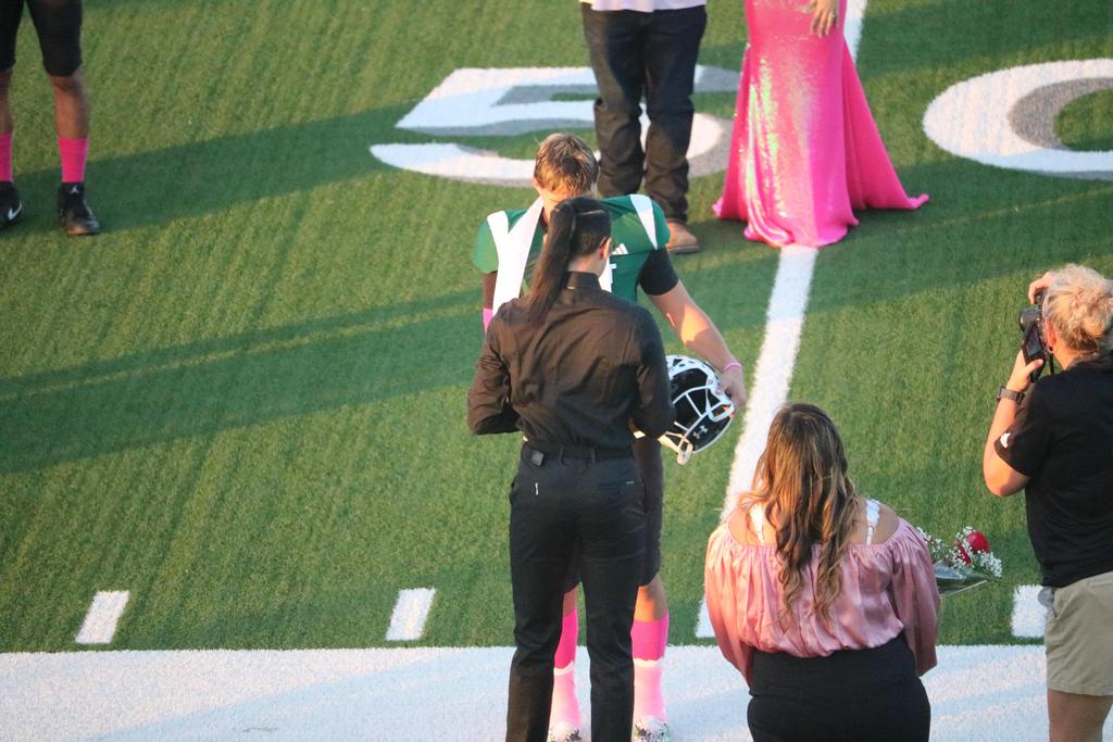 A woman in pink stands on a field alongside a football player, both engaged in a moment of camaraderie.