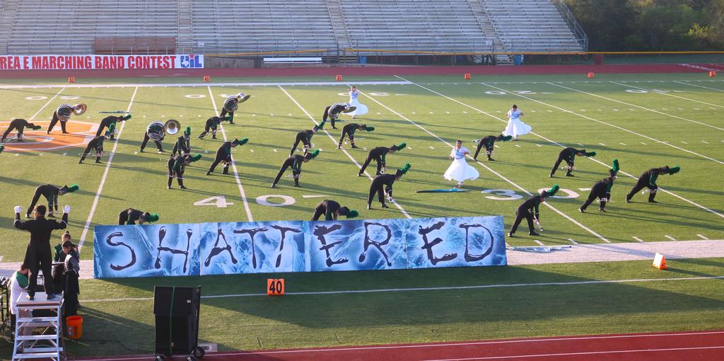 A marching band performs on a field, proudly displaying a banner that reads "Shattered" in bold letters.