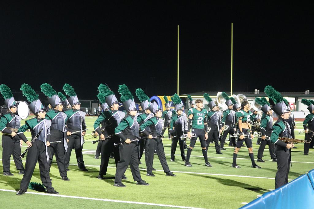 A vibrant marching band performs on a field under the night sky, illuminated by bright lights and surrounded by cheering spectators.