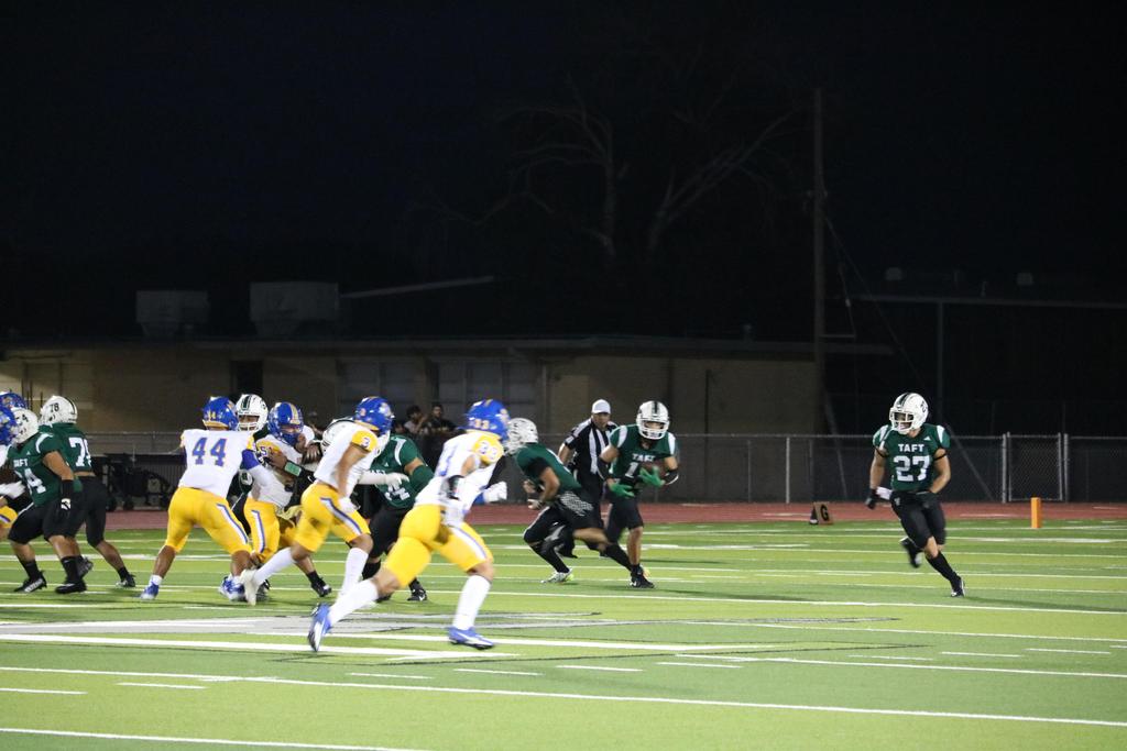 A night football game featuring players actively running and competing on the field under bright stadium lights.