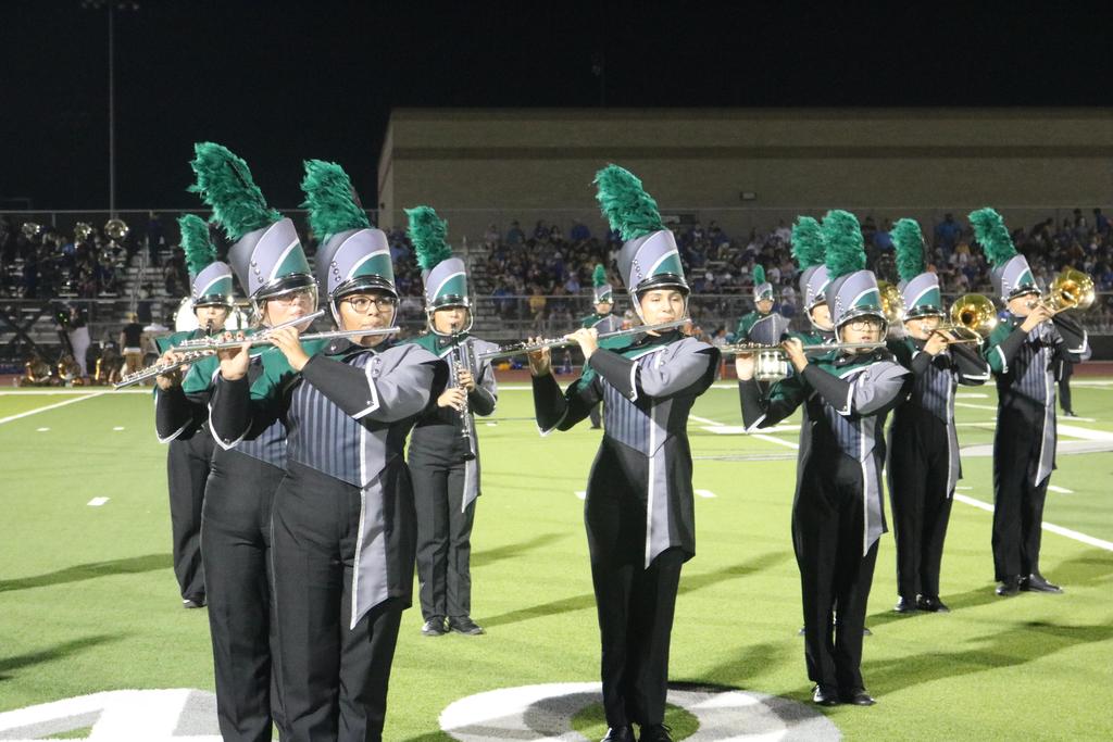 A vibrant marching band performs on a field under the night sky, illuminated by bright lights and surrounded by cheering spectators.