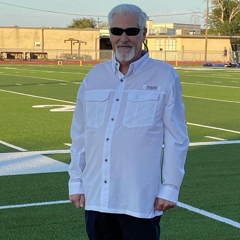 An older man stands confidently on a football field, surrounded by green grass and goalposts in the background.