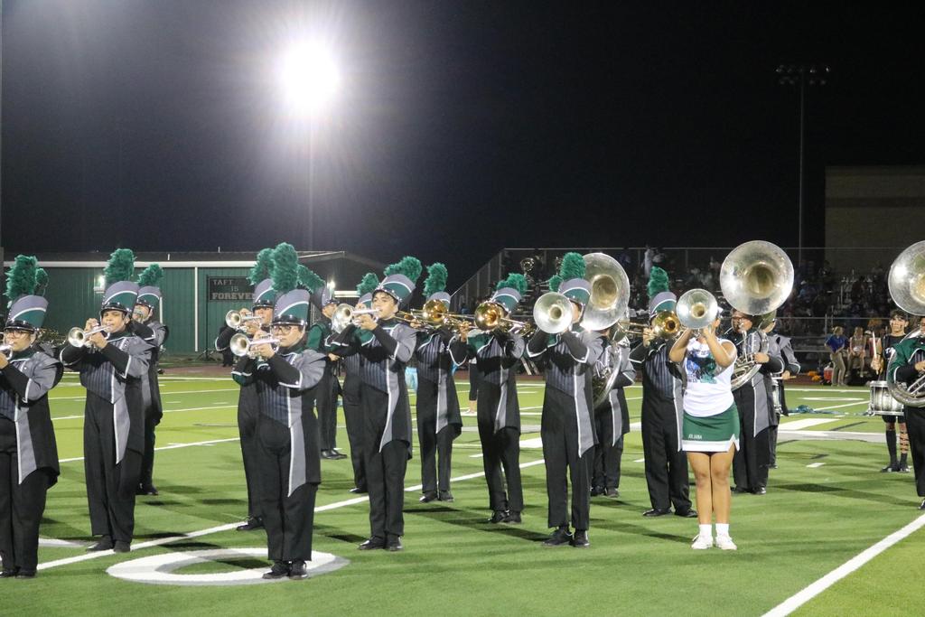 A vibrant marching band performs on a field under the night sky, illuminated by bright lights and surrounded by cheering spectators.