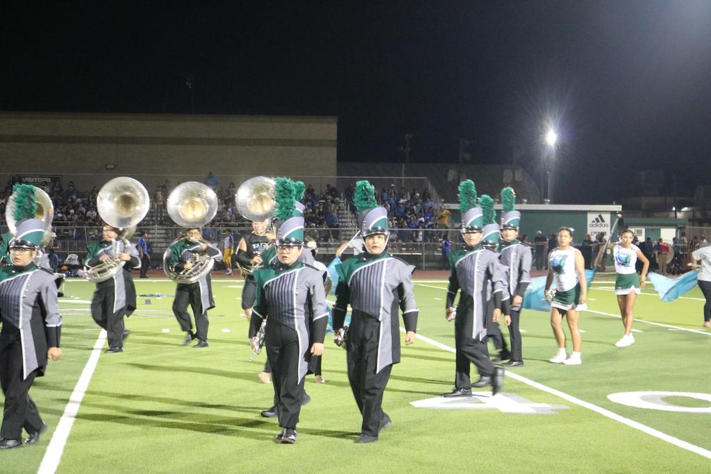 A vibrant marching band performs on a field under the night sky, illuminated by bright lights and surrounded by cheering spectators.