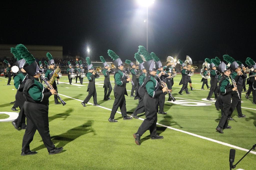 A marching band performs on a field at night, illuminated by bright lights, showcasing their uniforms and instruments.