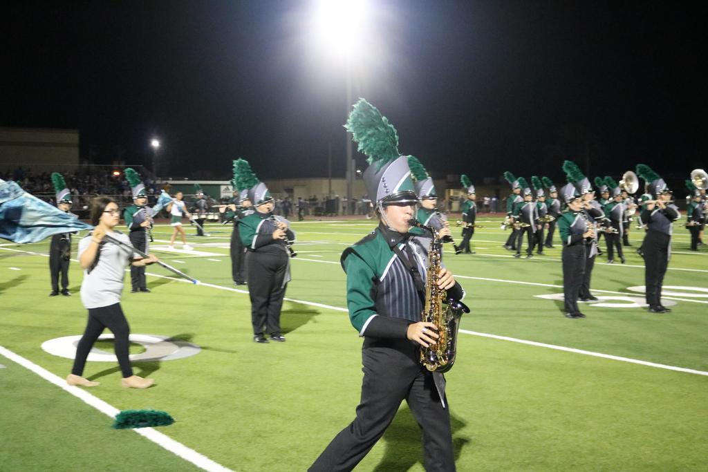 A vibrant marching band performs on a field under the night sky, illuminated by bright lights and surrounded by cheering spectators.