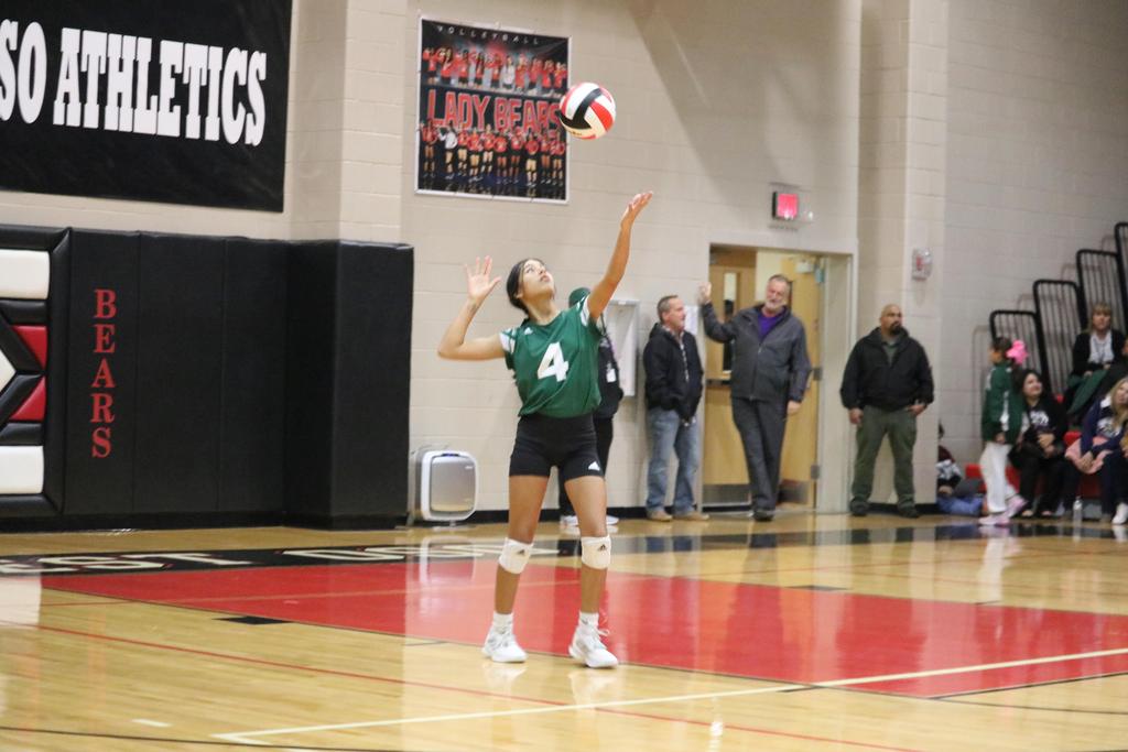 A girl in a green and black uniform skillfully serves a volleyball during a competitive match.