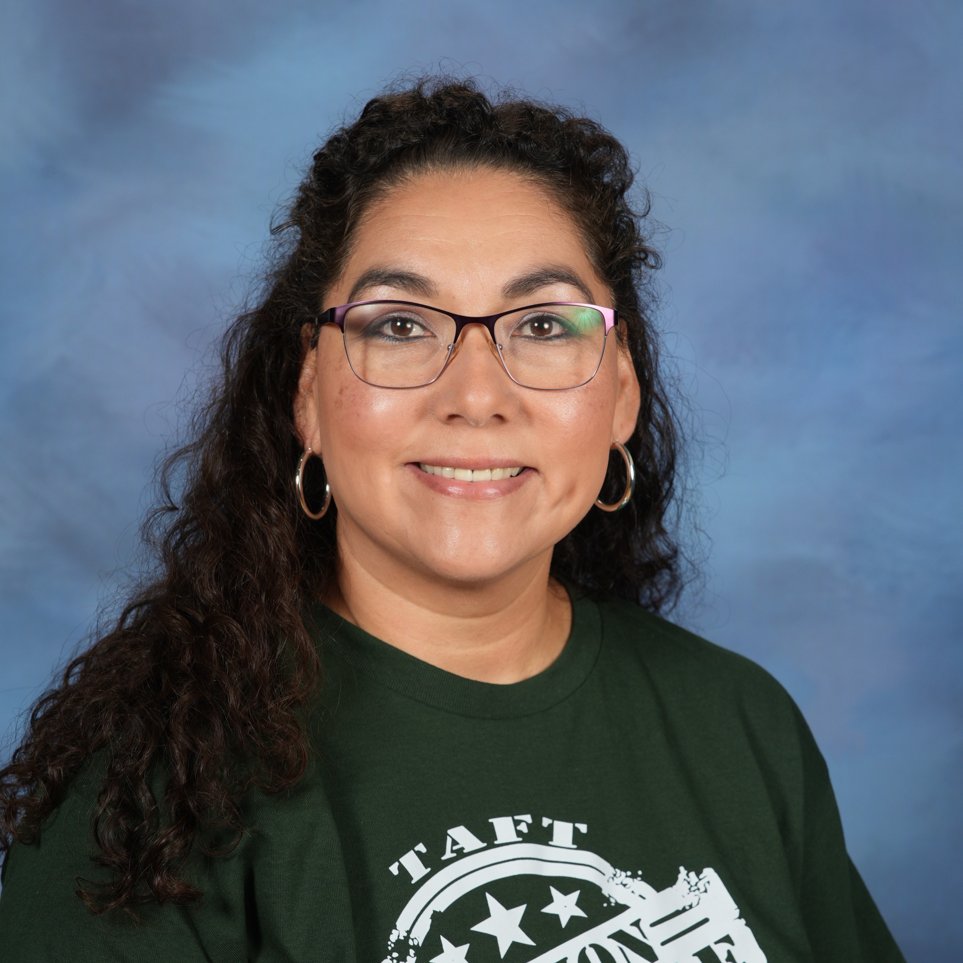 A woman wearing a green shirt and glasses, smiling while standing against a neutral background.