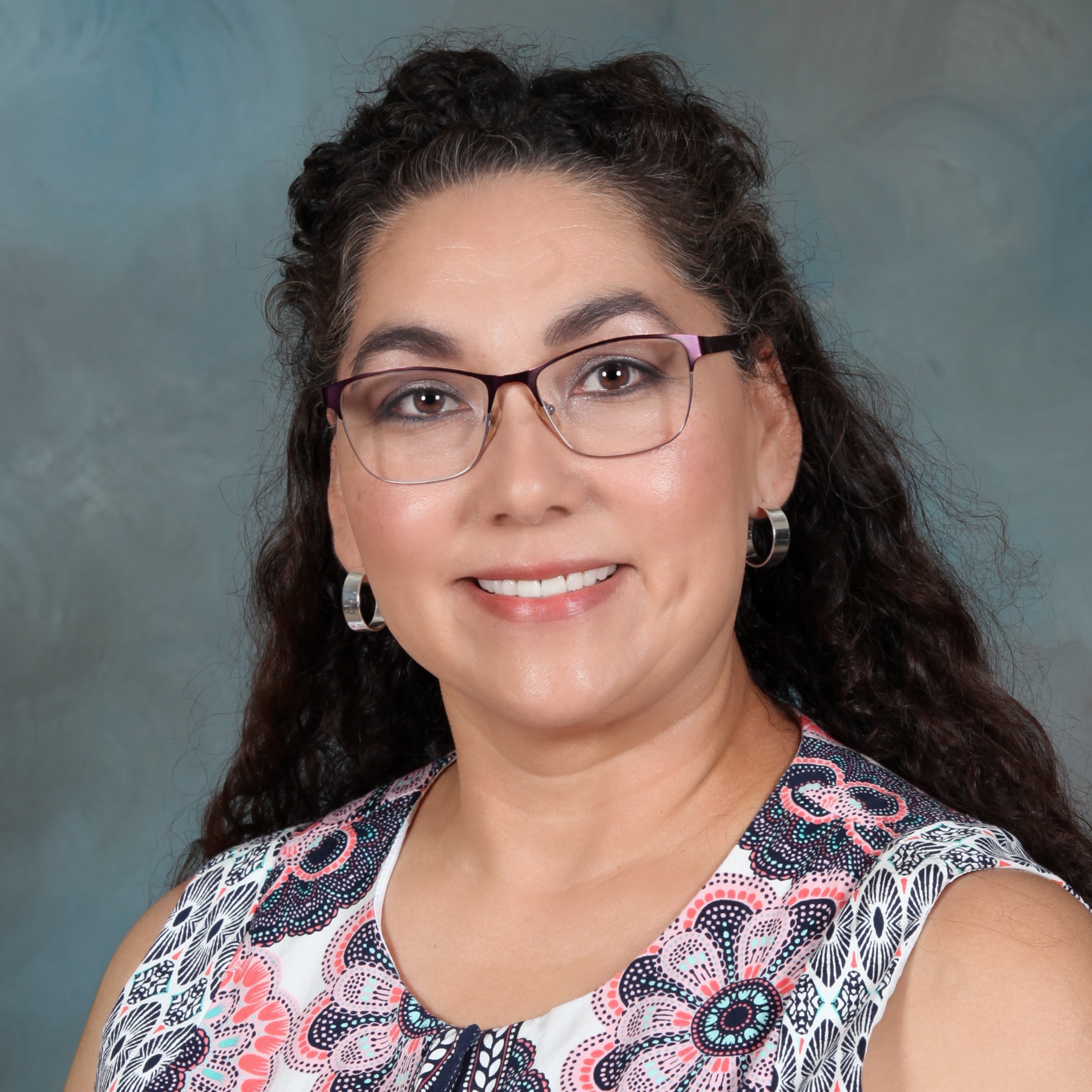 A woman with glasses wearing a grey shirt, looking confidently at the camera.