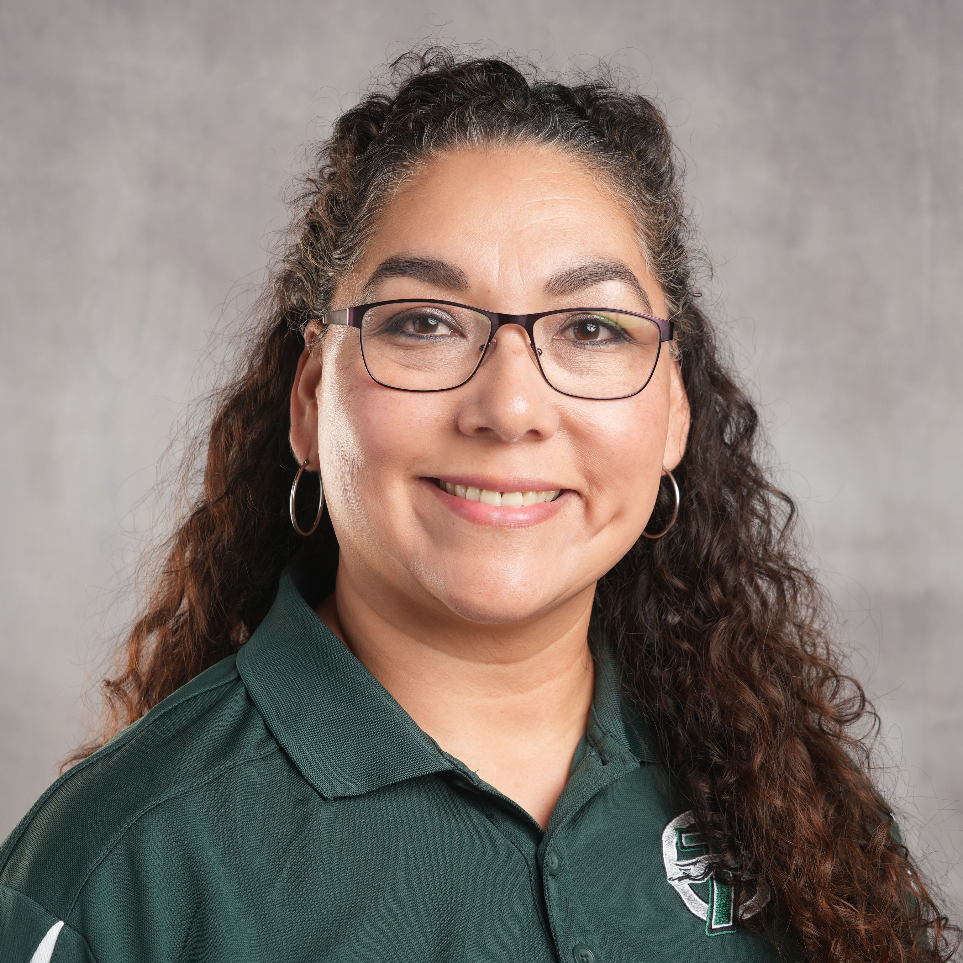 A woman wearing a green shirt and glasses, smiling while standing against a neutral background.