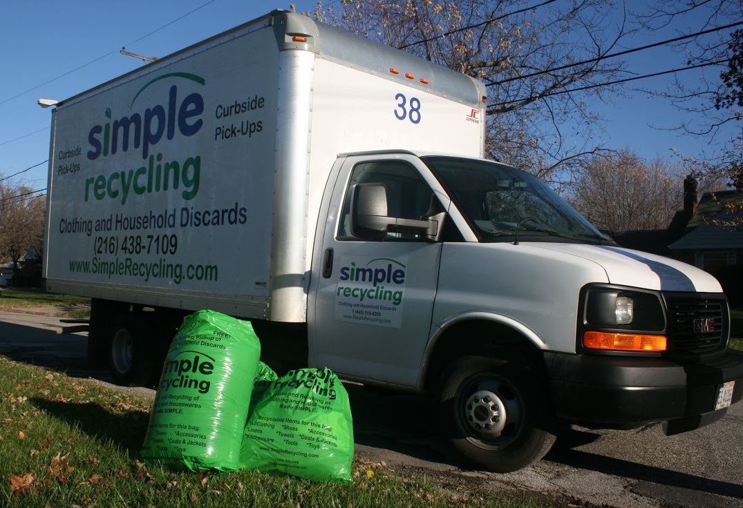 A white recycling truck labeled 'simple recycling' parked beside two green bags for collection on a residential street.