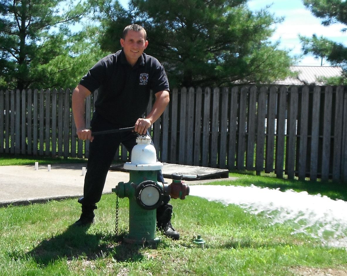 A person in a black shirt is operating a fire hydrant, with water flowing out onto the grass in a sunny outdoor setting.