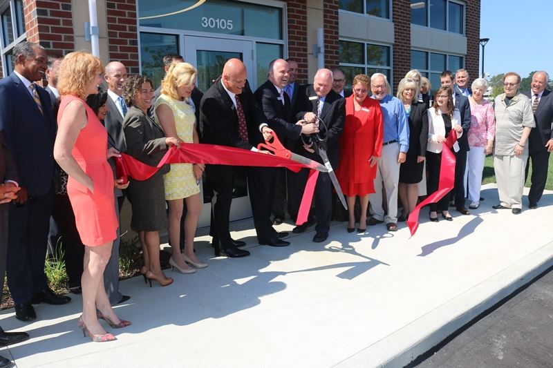 A group of people gathered outside a building, participating in a ribbon-cutting ceremony with a large red ribbon and scissors.