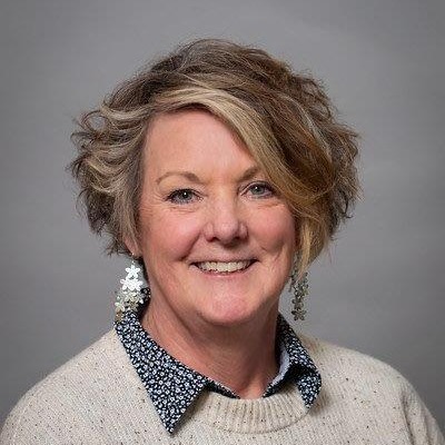 A woman with short, curly hair smiles at the camera, wearing a denim jacket over a pink collared shirt, set against a neutral gray background.