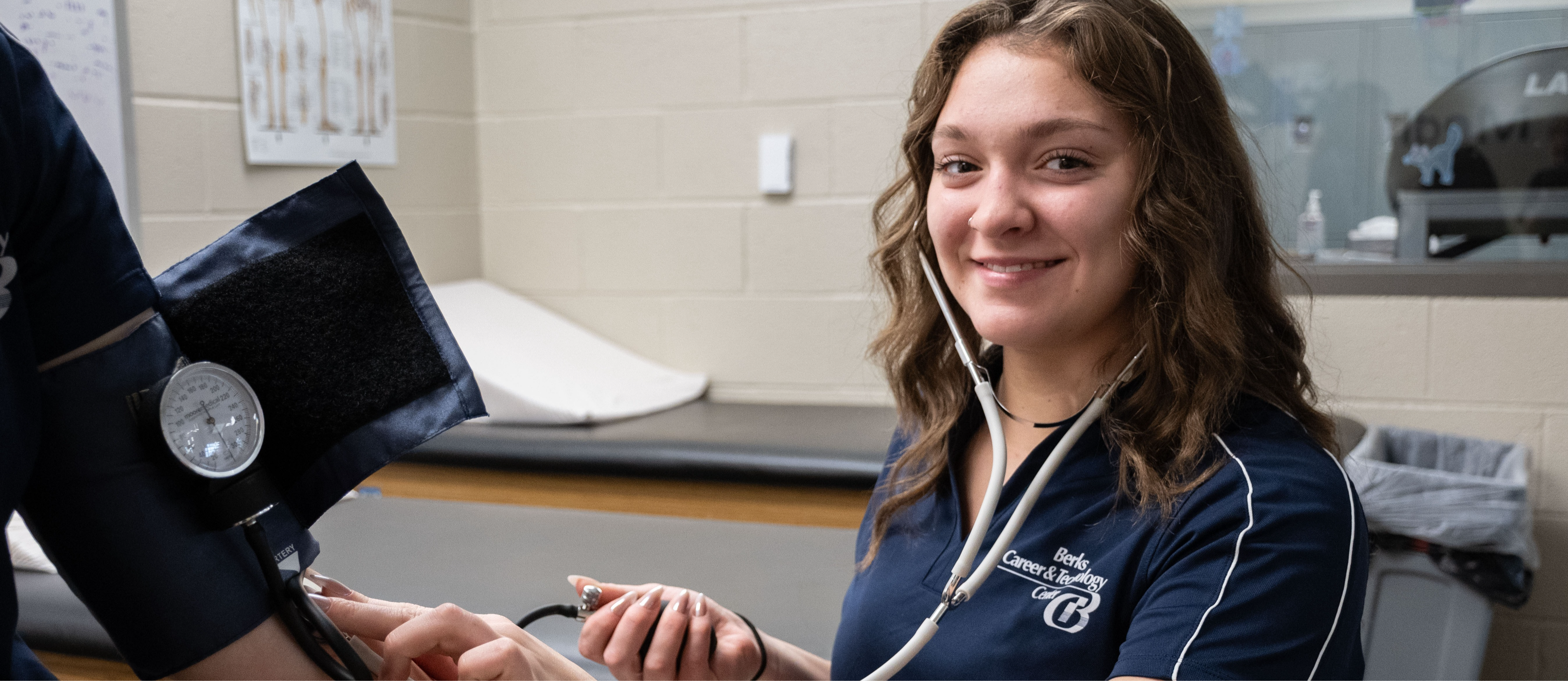 A student wearing a navy Berks Career & Technology Center polo shirt uses a stethoscope and blood pressure cuff to take another student’s blood pressure in a classroom lab setting. She smiles toward the camera while conducting the hands-on activity. The background includes exam tables, medical posters, and classroom equipment.