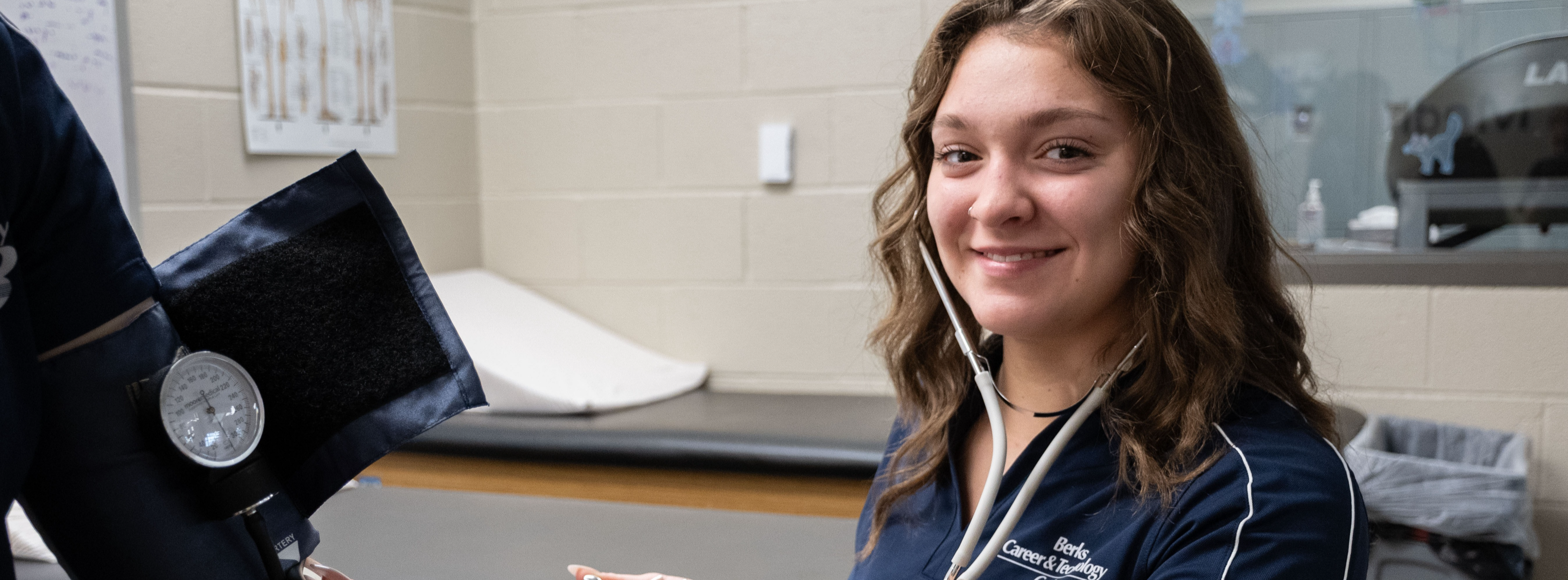 A student wearing a navy Berks Career & Technology Center polo shirt uses a stethoscope and blood pressure cuff to take another student’s blood pressure in a classroom lab setting. She smiles toward the camera while conducting the hands-on activity. The background includes exam tables, medical posters, and classroom equipment.