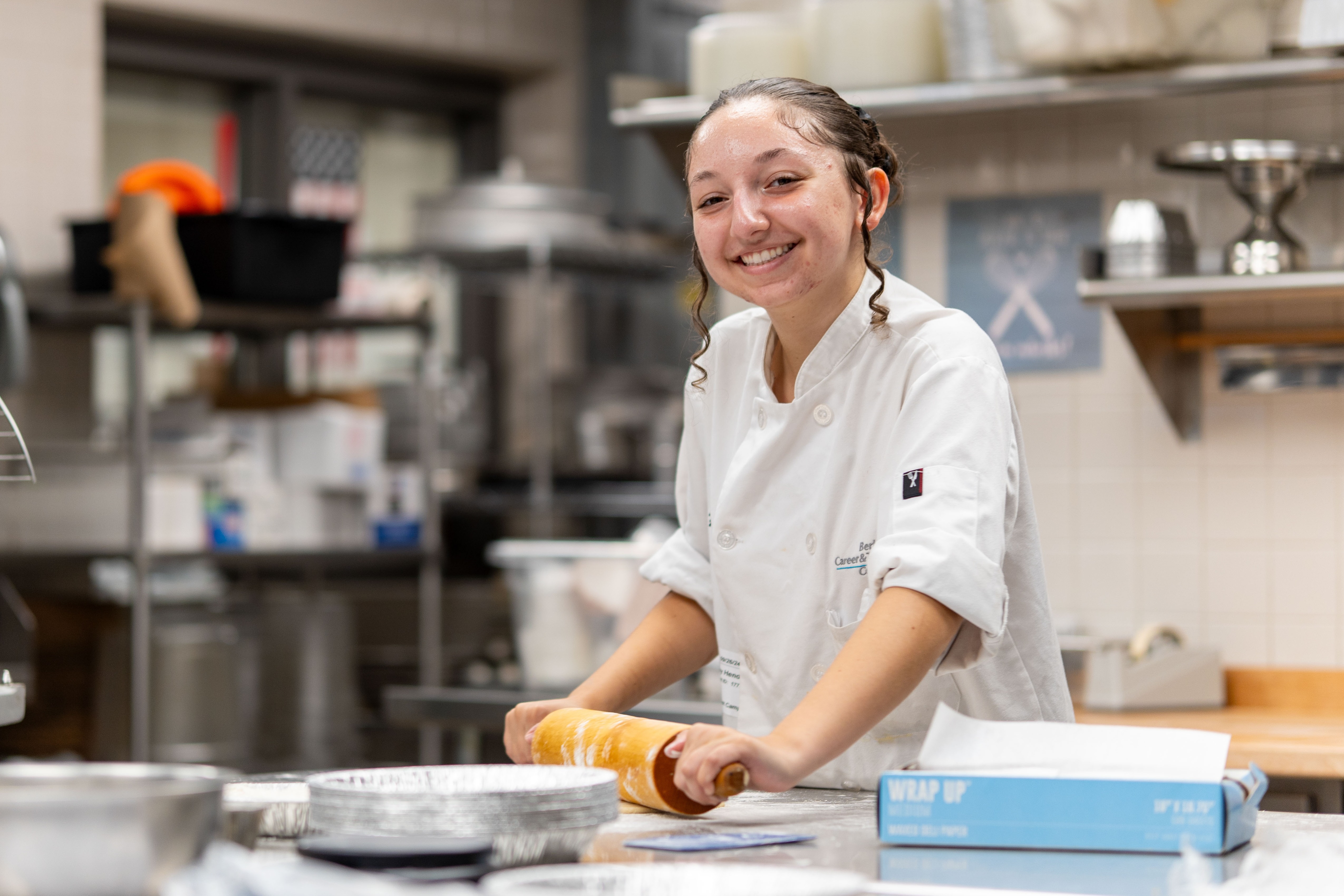 A culinary student in a white chef’s coat uses a rolling pin to prepare dough at a stainless-steel workstation in a commercial kitchen. Mixing bowls, baking pans, and kitchen equipment surround the workspace, and shelves with containers and supplies are visible in the background.