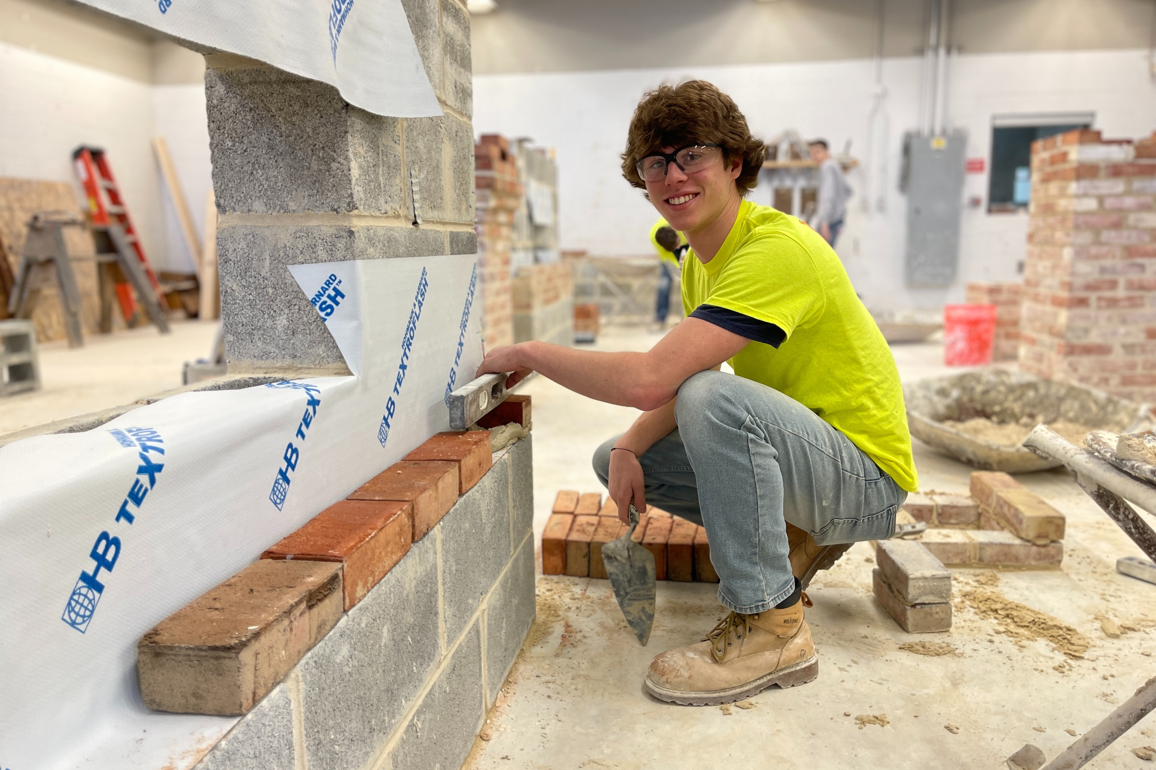A construction trainee wearing a bright yellow shirt and work boots is kneeling beside a partially built brick and concrete block wall. They are using a trowel to smooth mortar along the top row of bricks in an indoor workshop filled with masonry tools, materials, and other practice wall structures.