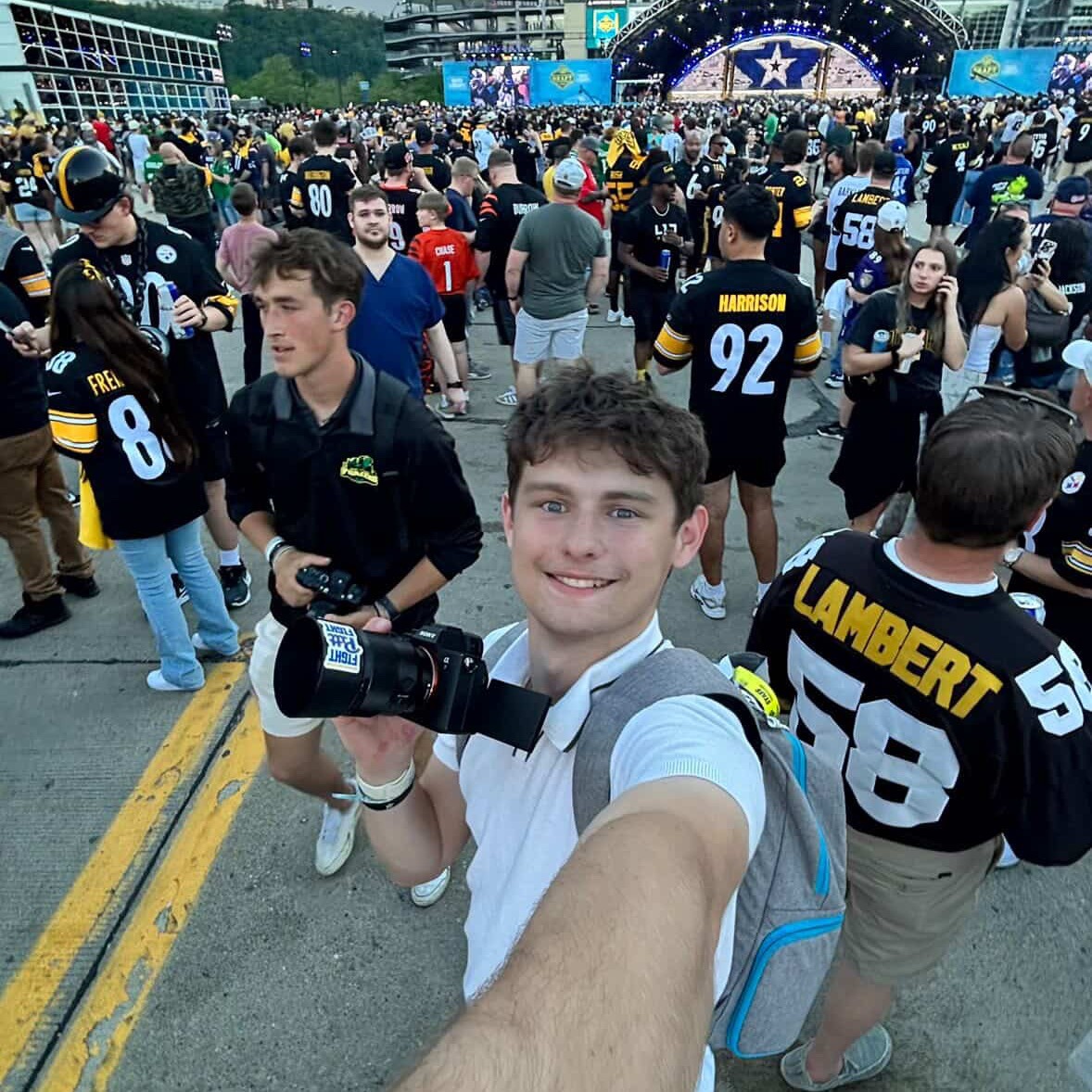 A crowd of people in sports jerseys gathers at a stadium. A man holds a camera taking a selfie.