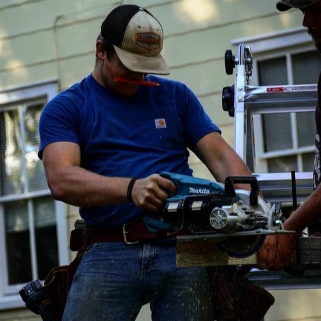Two men in blue shirts, one with a tool belt, hold a circular saw outside a house with white windows.