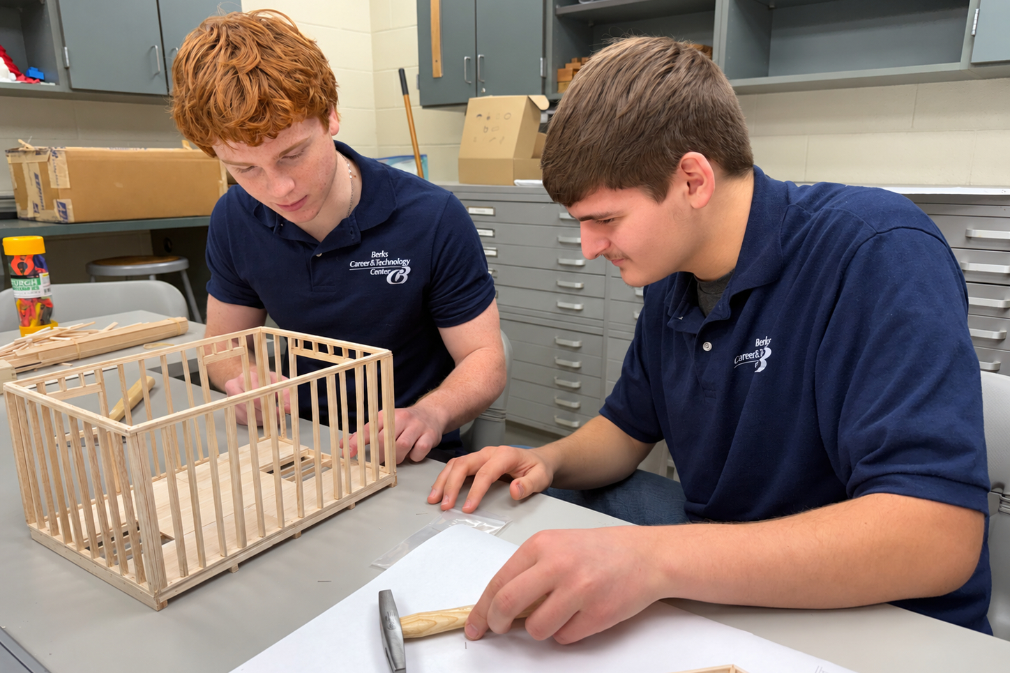 Two young men in blue polo shirts are seated at a table. One works on a wooden structure, while the other holds a hammer.