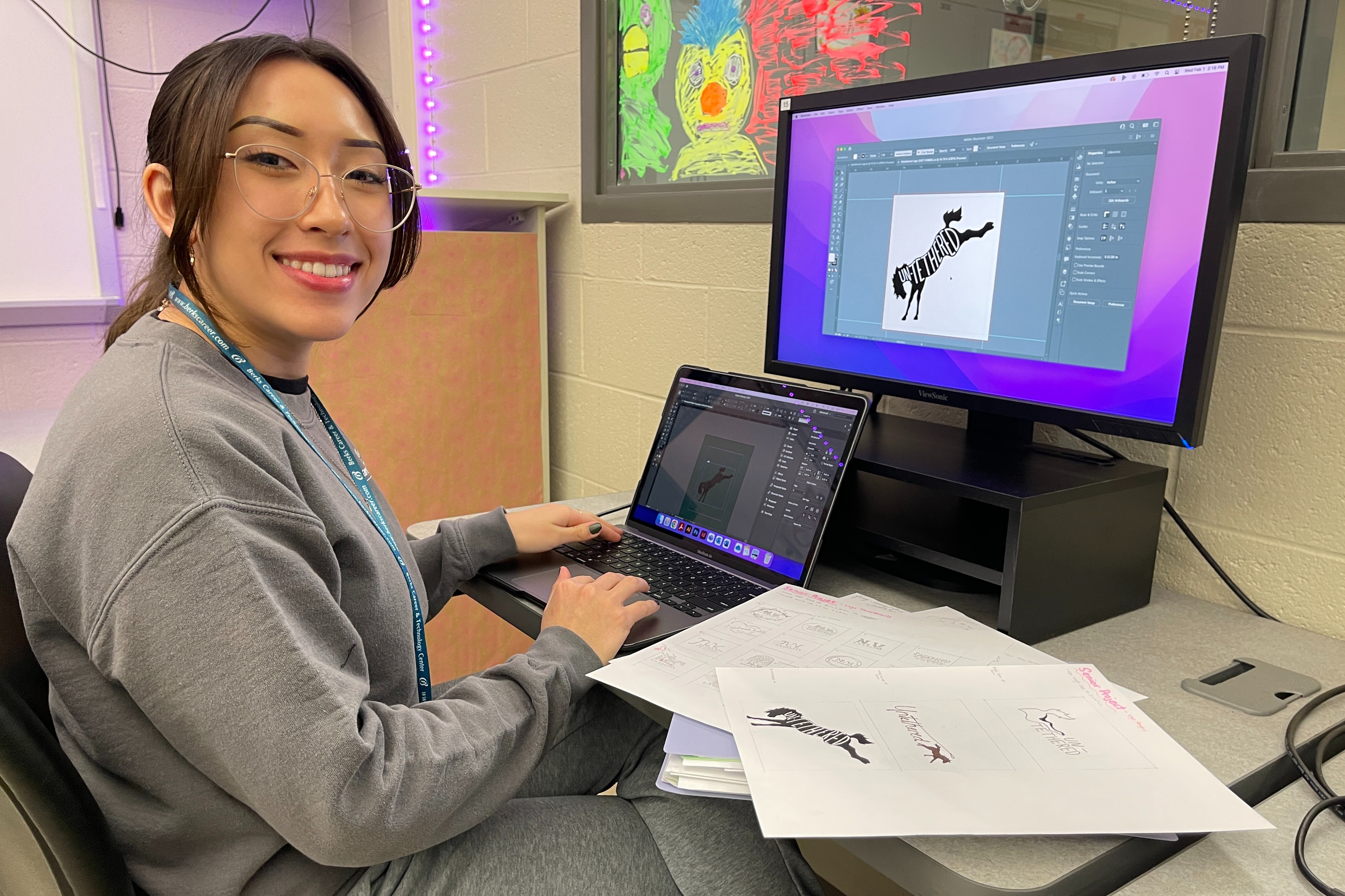 A person wearing glasses sits at a desk with a computer monitor and laptop, smiling.