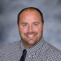 A man wearing a plaid shirt and tie smiles warmly at the camera.