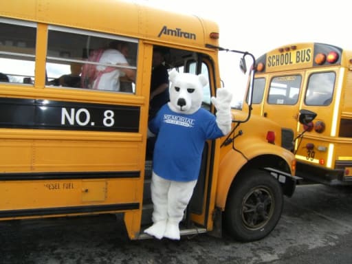 A person in a polar bear mascot costume waves from the doorway of a yellow school bus, with another bus visible in the background.