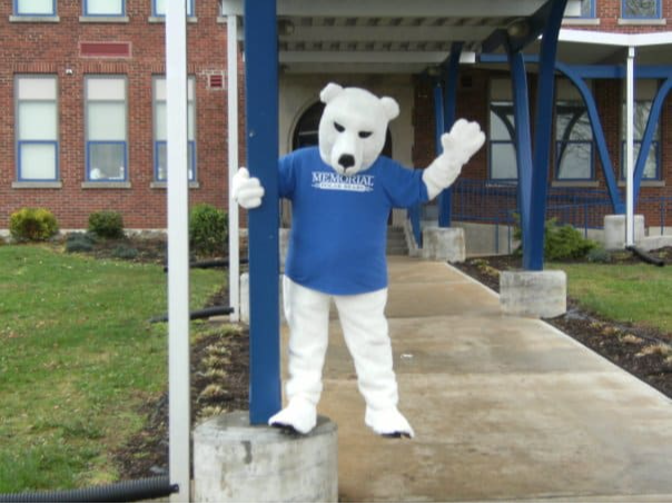 A polar bear mascot wearing a blue shirt waves from a sidewalk in front of a brick building.