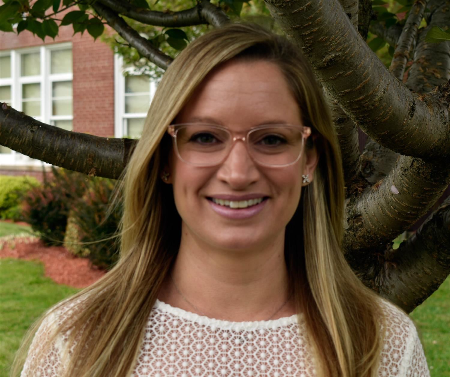 A woman with long blonde hair and glasses smiles in front of a tree, with a building visible in the background.