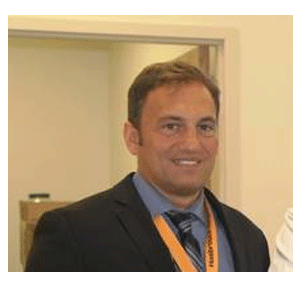 A man in a suit with a blue tie and an orange lanyard smiles while standing in a well-lit room.