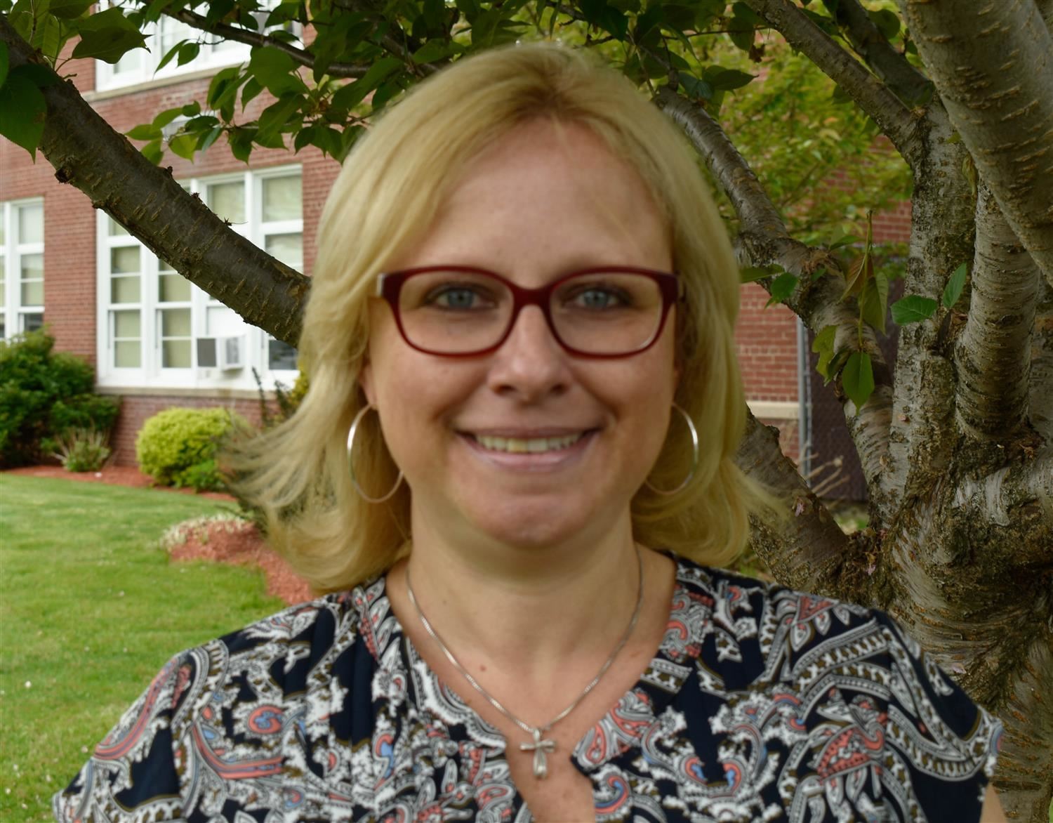 A smiling woman with blonde hair and glasses stands in front of a tree and a brick building, wearing a patterned blouse.