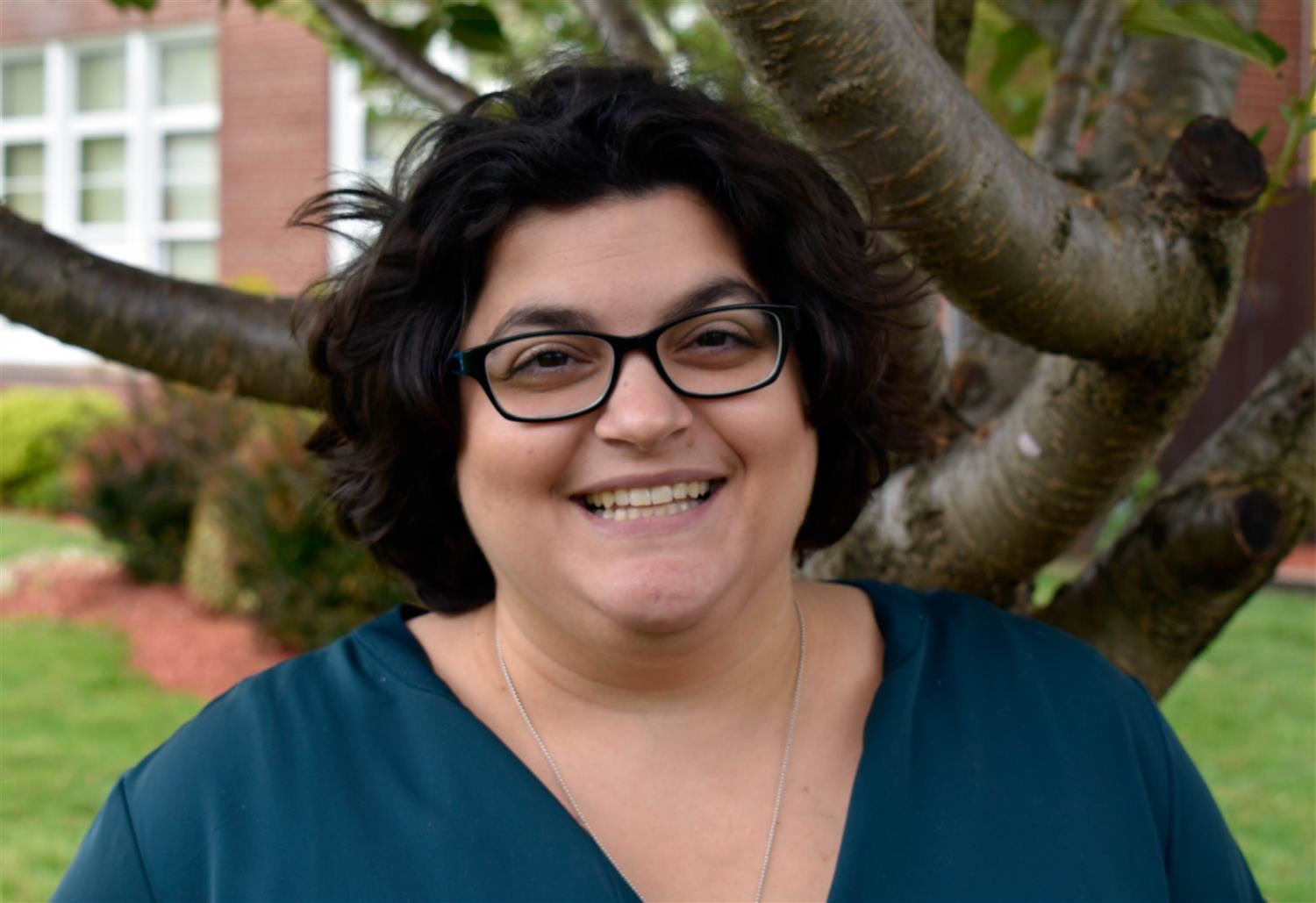 A smiling woman with curly dark hair and glasses stands in front of a tree, wearing a teal top, with a building visible in the background.