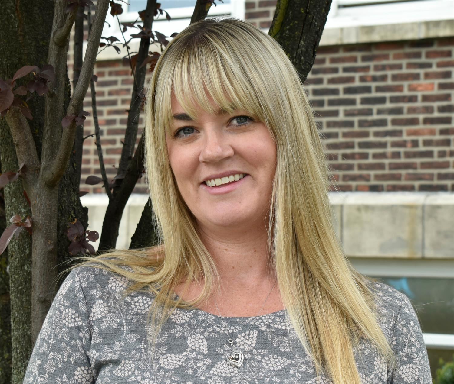 A woman with long blonde hair smiles while standing next to a tree, wearing a gray patterned shirt, with a brick building in the background.