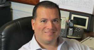 A man with short dark hair and a light-colored shirt smiles while seated in an office chair, with various office equipment visible in the background.