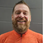 A bearded man wearing an orange shirt, standing confidently with a neutral expression.