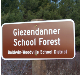 Sign for Giezendanner School Forest, located in Baldwin-Woodville School District, set against a backdrop of green trees.