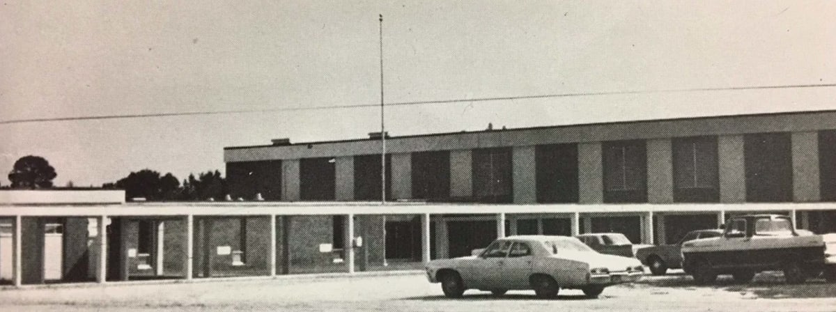 A black and white photograph of a mid-20th century building with a flat roof, featuring multiple windows and a parking lot filled with vintage cars.