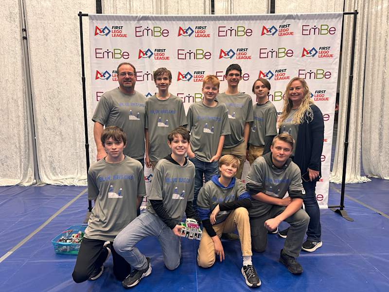 A group of ten individuals, wearing matching gray t-shirts, pose together in front of a branded backdrop at an event, with some members kneeling and others standing behind them.