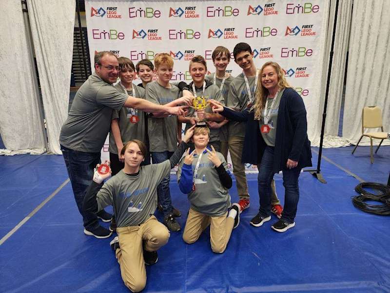 A group of people, including children and adults, celebrate together while holding a trophy in front of a backdrop with logos and event branding.