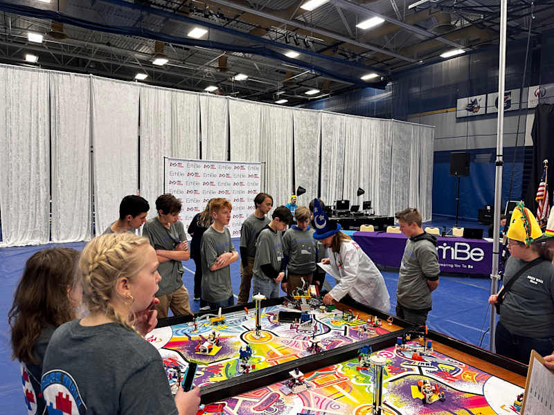 A group of students in gray shirts are gathered around a colorful robotics competition table, with various robots and equipment, in a gymnasium setting.