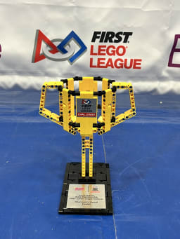 A trophy made of yellow and black LEGO bricks, featuring a plaque at the base, displayed on a blue surface with a backdrop of a FIRST LEGO League banner.