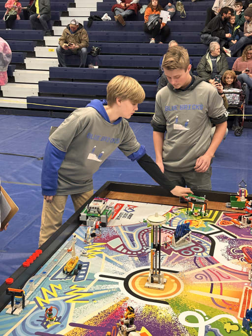 Two young boys in gray team shirts are focused on a colorful robotics competition table, with various models and structures set up for their project, while spectators watch from the bleachers in the background.