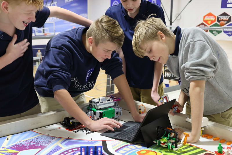 A group of four students, two boys and two girls, are gathered around a laptop on a table, engaged in a robotics project, with various robotic components and a colorful mat in the background.
