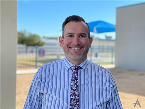 A smiling man in a striped shirt and floral tie stands outdoors with a blue sky and a shaded area in the background.