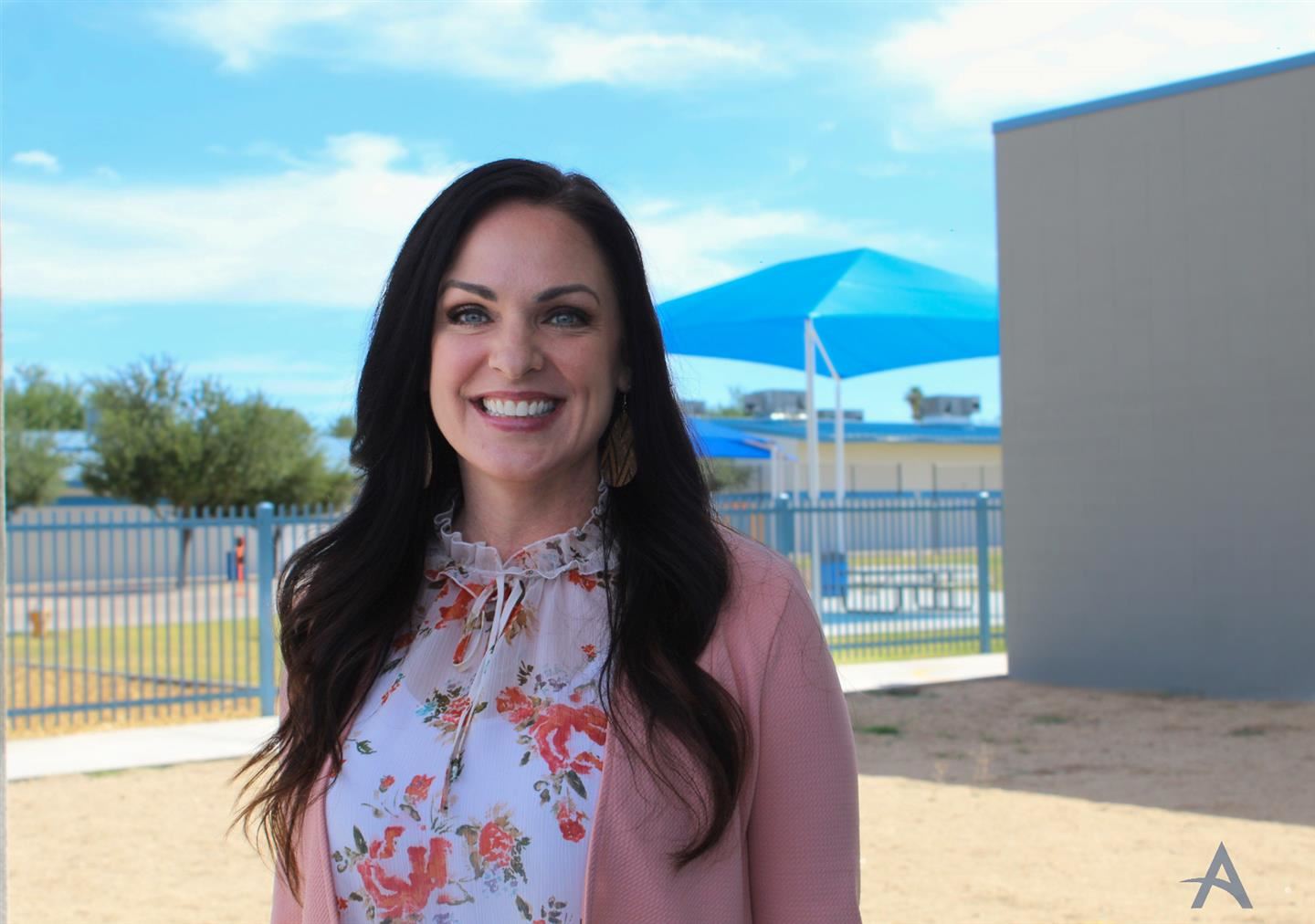 A woman with long dark hair smiles in front of a school playground, wearing a floral blouse and a pink blazer, with blue shade structures in the background.