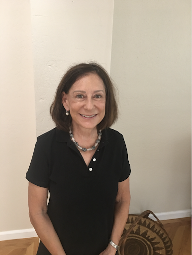 A woman with shoulder-length brown hair, wearing a black polo shirt and a necklace, smiles while standing in a well-lit room with wooden flooring and a decorative basket in the background.
