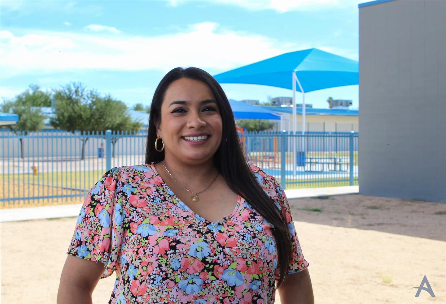 A woman with long dark hair smiles while standing outdoors in front of a school playground, wearing a colorful floral dress, with blue canopies and a clear sky in the background.